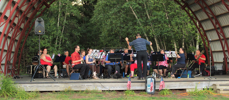 Picture of the band performing outdoors under a performance shell with trees behind them